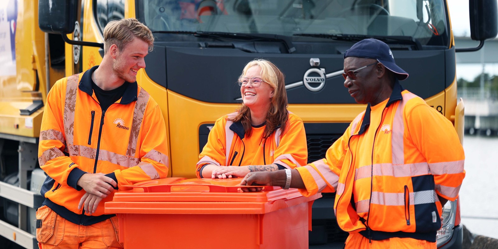 Medarbetare står och lutar sig mot orange kärl framför en baklastare (sopbil)